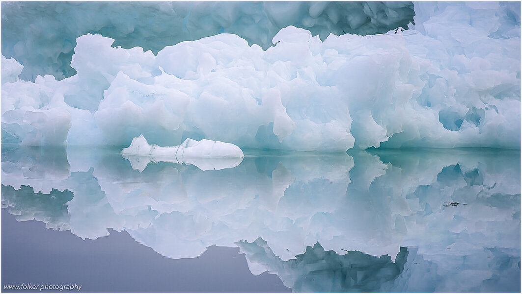 The backdrop of this fine-art photograph is formed and filled by a bigger iceberg with reflection in the water. Smaller elegantly shaped pieces of ice are passing in the foreground. Different shades of ice-blue color tones and cracks in the ice create a symmetric and well-balanced image that almost feels abstract. Picture was taken at the Sermilik fjord in East-Greenland.
