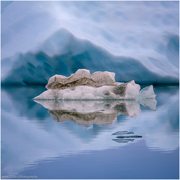 The backdrop of this fine-art photograph is formed and filled by a bigger iceberg with reflection in the water. Smaller elegantly shaped pieces of ice are passing in the foreground. Different shades of ice-blue color tones and cracks in the ice create a symmetric and well-balanced image that almost feels abstract. Picture was taken at the Sermilik fjord in East-Greenland.