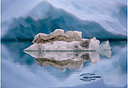 The backdrop of this fine-art photograph is formed and filled by a bigger iceberg with reflection in the water. Smaller elegantly shaped pieces of ice are passing in the foreground. Different shades of ice-blue color tones and cracks in the ice create a symmetric and well-balanced image that almost feels abstract. Picture was taken at the Sermilik fjord in East-Greenland.