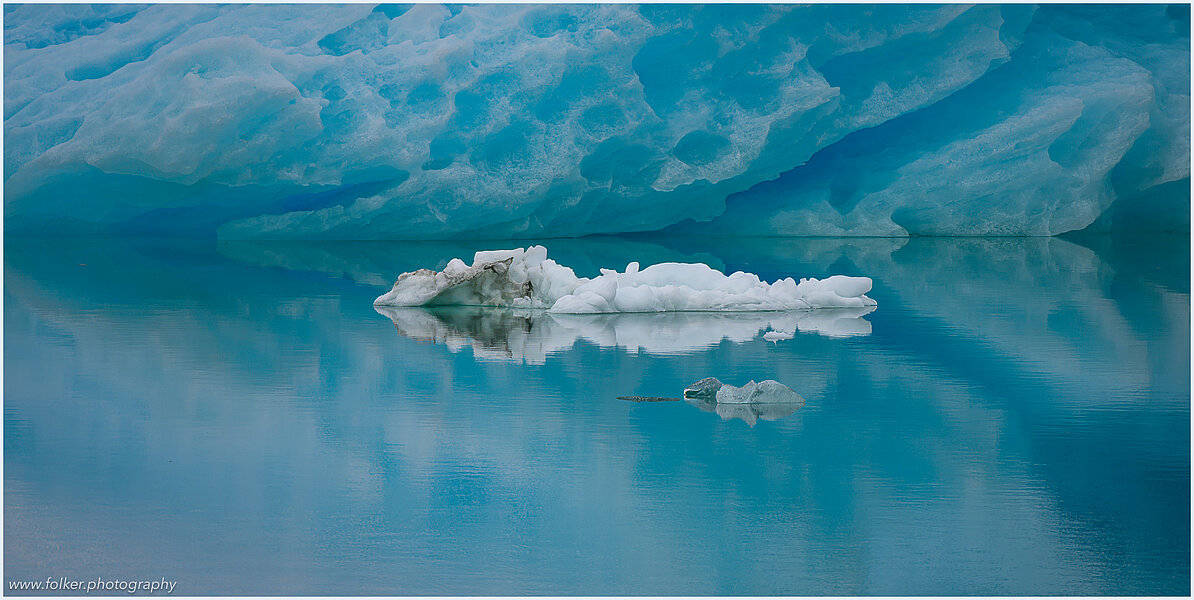 The backdrop of this fine-art photograph is formed and filled by a bigger iceberg with reflection in the water. Smaller elegantly shaped pieces of ice are passing in the foreground. Different shades of ice-blue color tones and cracks in the ice create a symmetric and well-balanced image that almost feels abstract. Picture was taken at the Sermilik fjord in East-Greenland.