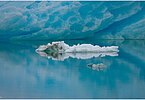 The backdrop of this fine-art photograph is formed and filled by a bigger iceberg with reflection in the water. Smaller elegantly shaped pieces of ice are passing in the foreground. Different shades of ice-blue color tones and cracks in the ice create a symmetric and well-balanced image that almost feels abstract. Picture was taken at the Sermilik fjord in East-Greenland.