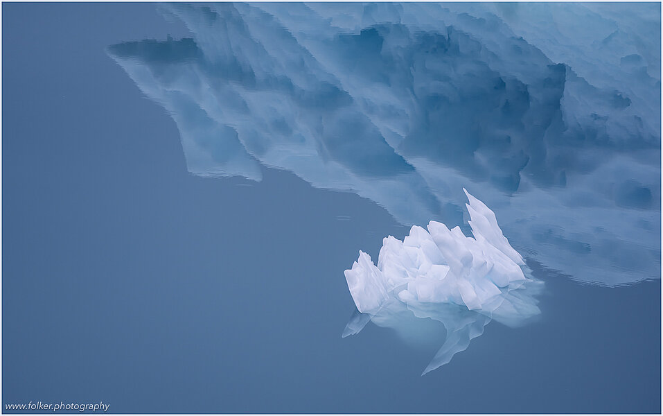 This fine-art photograph shows a minimalistic detail of a small iceberg in front of the reflection of a bigger iceberg. Picture was taken at the Sermilik fjord in East-Greenland.