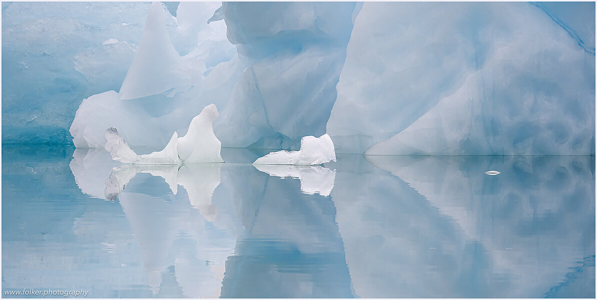 The backdrop of this fine-art photograph is formed and filled by a bigger iceberg with reflection in the water. Smaller elegantly shaped pieces of ice are passing in the foreground. Different shades of ice-blue color tones and cracks in the ice create a symmetric and well-balanced image that almost feels abstract. Picture was taken at the Sermilik fjord in East-Greenland.