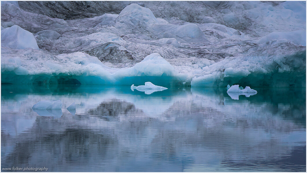 The backdrop of this fine-art photograph is formed and filled by a bigger iceberg with reflection in the water. Smaller elegantly shaped pieces of ice are passing in the foreground. Different shades of ice-blue color tones and cracks in the ice create a symmetric and well-balanced image that almost feels abstract. Picture was taken at the Sermilik fjord in East-Greenland.