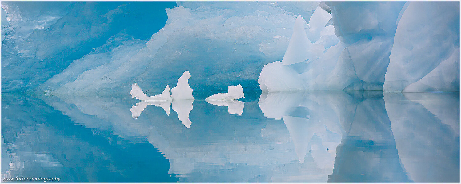 The backdrop of this fine-art photograph is formed and filled by a bigger iceberg with reflection in the water. Smaller elegantly shaped pieces of ice are passing in the foreground. Different shades of ice-blue color tones and cracks in the ice create a symmetric and well-balanced image that almost feels abstract. Picture was taken at the Sermilik fjord in East-Greenland. This image has an unusual aspect ratio of 1:3.