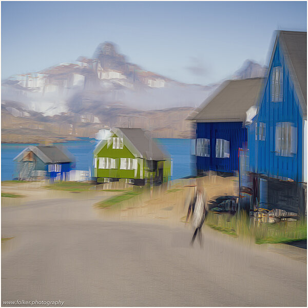 This fine-art photograph shows an abstract, painterly street scene in Tassilaq in East-Greenland. In a style of a painting from early 20th century, you can see people walking down a road towards the fjord with colorful houses, mountains and clouds in the backdrop. 