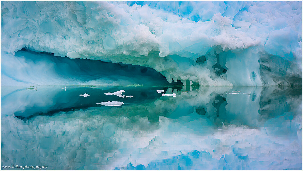 The backdrop of this fine-art photograph is formed and filled by a bigger iceberg with reflection in the water. Smaller elegantly shaped pieces of ice are passing in the foreground. Different shades of ice-blue color tones and cracks in the ice create a symmetric and well-balanced image that almost feels abstract. Picture was taken at the Sermilik fjord in East-Greenland.