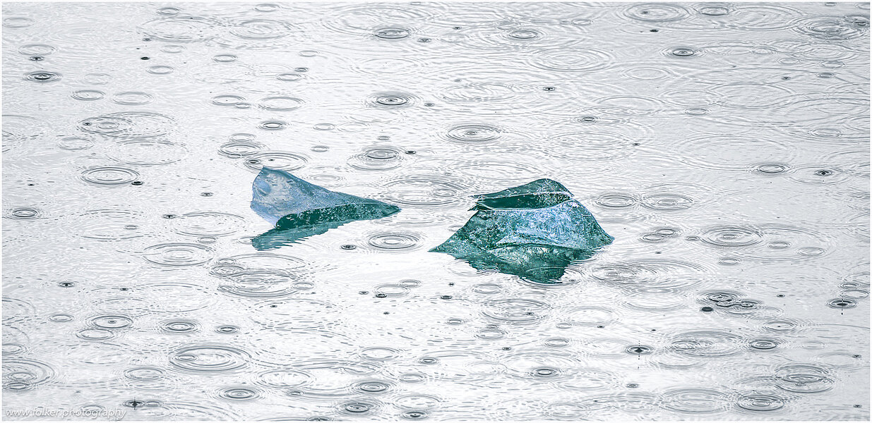 This fine-art photograph showing a minimalistic detail of a small iceberg surrounded by water drops from rain. Picture was taken at the Sermilik fjord in East-Greenland.