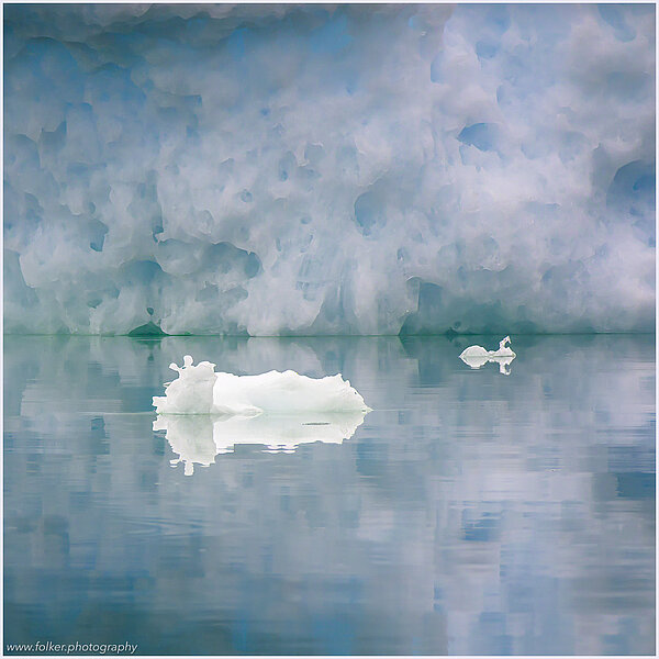 The backdrop of this fine-art photograph is formed and filled by a bigger iceberg with reflection in the water. Smaller elegantly shaped pieces of ice are passing in the foreground. Different shades of ice-blue color tones and cracks in the ice create a symmetric and well-balanced image that almost feels abstract. Picture was taken at the Sermilik fjord in East-Greenland.