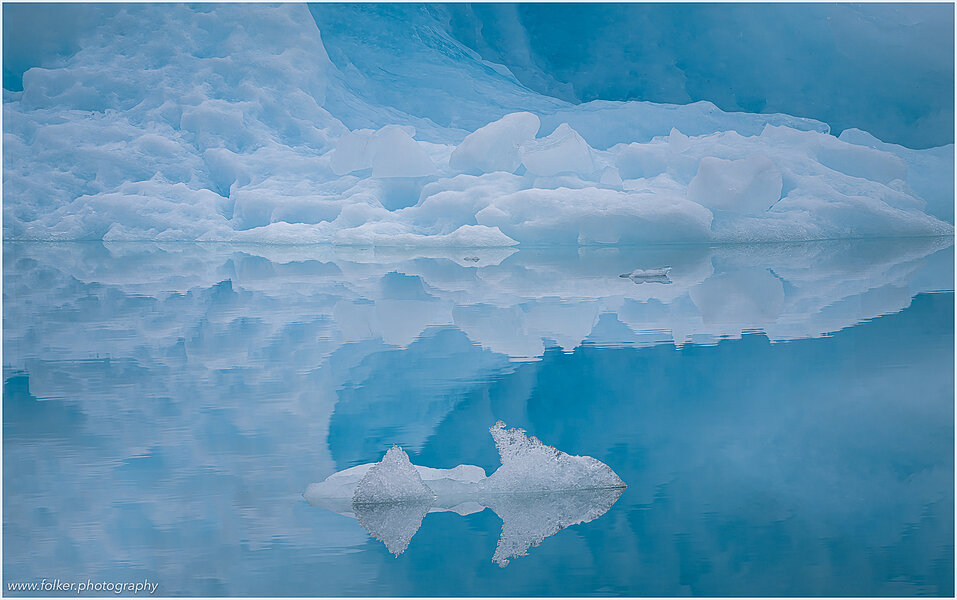 The backdrop of this fine-art photograph is formed and filled by a bigger iceberg with reflection in the water. Smaller elegantly shaped pieces of ice are passing in the foreground. Different shades of ice-blue color tones and cracks in the ice create a symmetric and well-balanced image that almost feels abstract. Picture was taken at the Sermilik fjord in East-Greenland.
