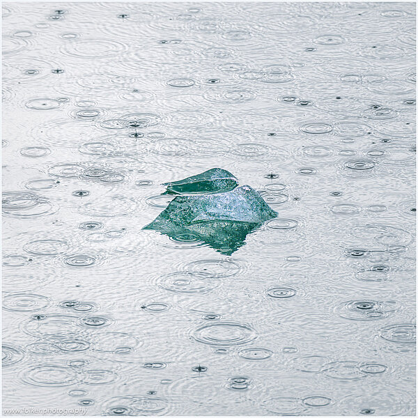 This fine-art photograph showing a minimalistic detail of a small iceberg surrounded by water drops from rain. Picture was taken at the Sermilik fjord in East-Greenland.