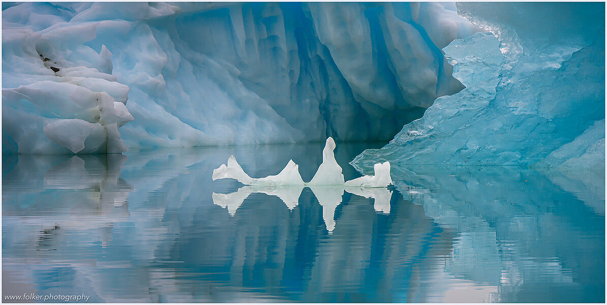The backdrop of this fine-art photograph is formed and filled by a bigger iceberg with reflection in the water. Smaller elegantly shaped pieces of ice are passing in the foreground. Different shades of ice-blue color tones and cracks in the ice create a symmetric and well-balanced image that almost feels abstract. Picture was taken at the Sermilik fjord in East-Greenland.