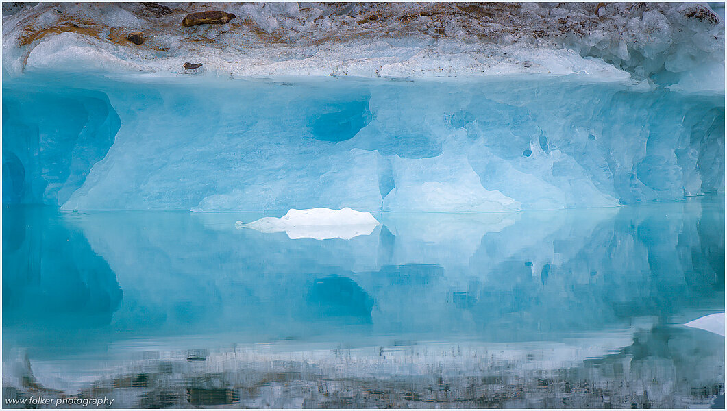 The backdrop of this fine-art photograph is formed and filled by a bigger iceberg with reflection in the water. Smaller elegantly shaped pieces of ice are passing in the foreground. Different shades of ice-blue color tones and cracks in the ice create a symmetric and well-balanced image that almost feels abstract. Picture was taken at the Sermilik fjord in East-Greenland.