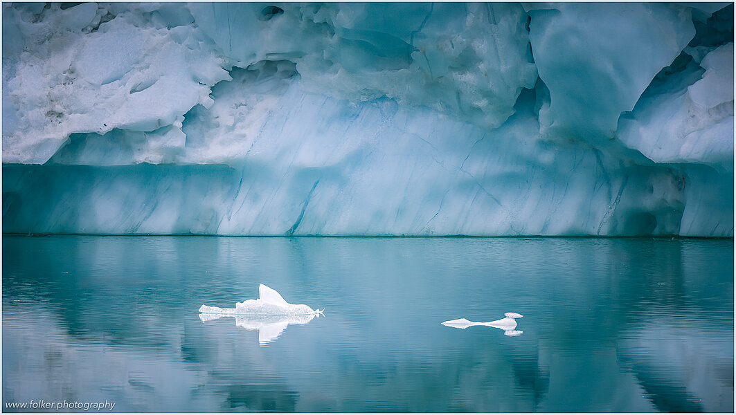 The backdrop of this fine-art photograph is formed and filled by a bigger iceberg with reflection in the water. Smaller elegantly shaped pieces of ice are passing in the foreground. Different shades of ice-blue color tones and cracks in the ice create a symmetric and well-balanced image that almost feels abstract. Picture was taken at the Sermilik fjord in East-Greenland.