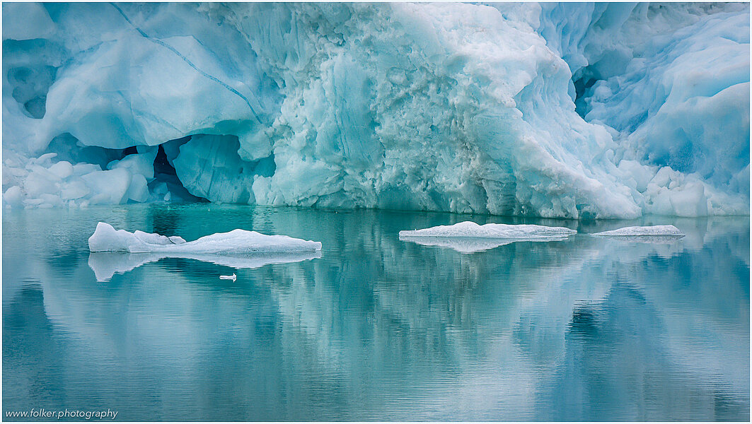 The backdrop of this fine-art photograph is formed and filled by a bigger iceberg with reflection in the water. Smaller elegantly shaped pieces of ice are passing in the foreground. Different shades of ice-blue and green color tones and cracks in the ice create a symmetric and well-balanced image that almost feels abstract. Picture was taken at the Sermilik fjord in East-Greenland.