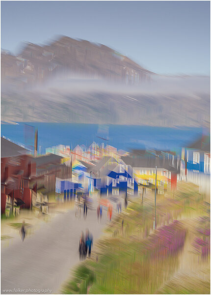 This fine-art photograph shows an abstract, painterly street scene in Tassilaq in East-Greenland. In a style of a painting from early 20th century, you can see people walking down a road towards the fjord with mountains and clouds in the backdrop. 