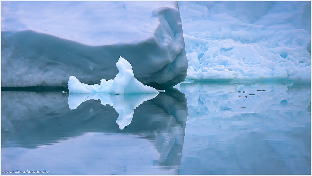 The backdrop of this fine-art photograph is formed and filled by a bigger iceberg with reflection in the water. Smaller elegantly shaped pieces of ice are passing in the foreground. Different shades of ice-blue color tones and cracks in the ice create a symmetric and well-balanced image that almost feels abstract. Picture was taken at the Sermilik fjord in East-Greenland.