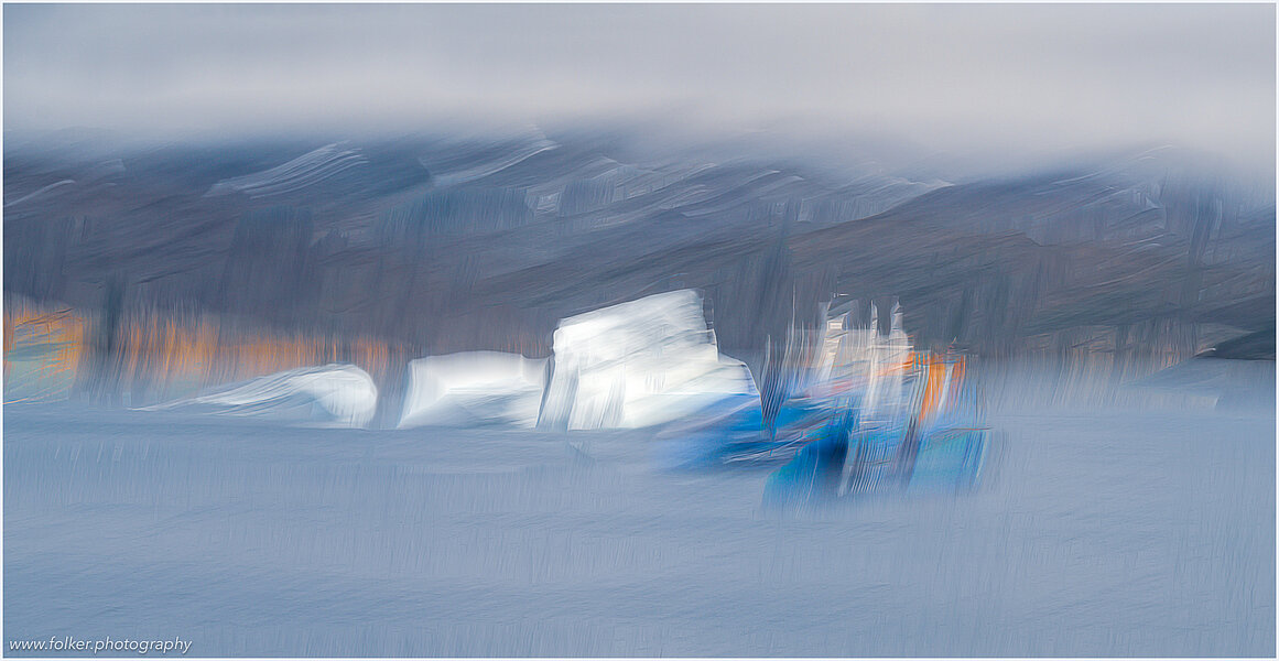 This fine-art photograph shows an abstract, painterly scene in Tassilaq in East-Greenland. In a style of a painting from early 20th century, you can imagine to see an iceberg and a blue ferry in front of cloudy mountains.